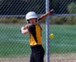 Softball player in yellow jersey preparing to hit a pitch on a sunny day near chain-link fence.