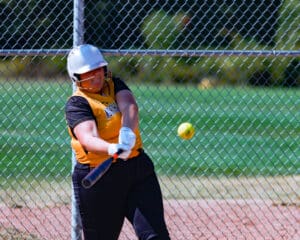 Softball player in yellow jersey hitting a ball during a game, with a chain-link fence in the background.