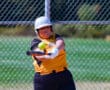Softball player in yellow jersey hitting a ball with a bat near a chain-link fence on a sunny day.