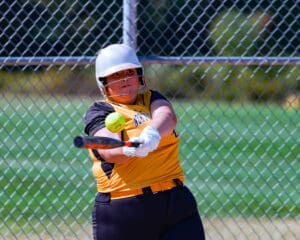 Softball player in yellow jersey hitting a ball with a bat near a chain-link fence on a sunny day.
