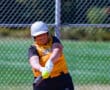 Softball player in action, swinging bat, yellow jersey, helmet on, against chain-link fence background.