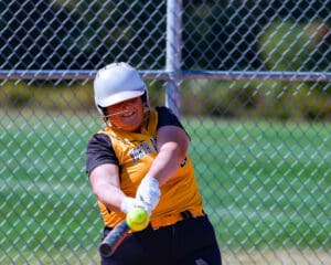 Softball player in action, swinging bat, yellow jersey, helmet on, against chain-link fence background.