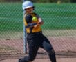 Softball player in yellow and black uniform swinging bat, hitting ball at a game, with fence and field in background.