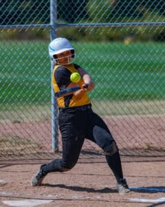 Softball player in yellow and black uniform swinging bat, hitting ball at a game, with fence and field in background.