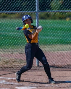 Softball player in action swinging a bat, hitting a ball on a sunny day at the field.