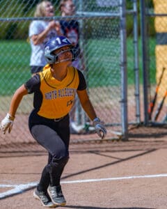 Softball player in yellow jersey and helmet prepares to run at a game, with spectators in the background.