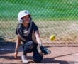 Catcher in protective gear ready to catch a softball on the field, with a chain-link fence background.
