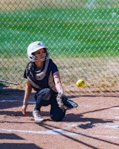 Catcher in protective gear ready to catch a softball on the field, with a chain-link fence background.
