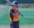 Softball player ready to bat, wearing a yellow jersey and helmet, standing near a chain-link fence on a sunny day.