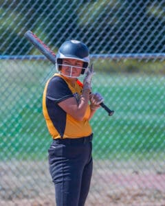 Softball player ready to bat, wearing a yellow jersey and helmet, standing near a chain-link fence on a sunny day.