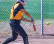 Softball player in yellow batting during a game, wearing a helmet and black pants, swings as the ball approaches.