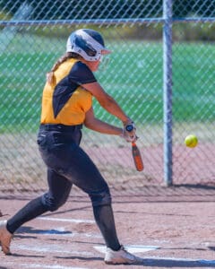 Softball player in yellow batting during a game, wearing a helmet and black pants, swings as the ball approaches.