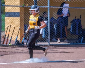 Softball player in action, crossing home plate on a sunny day, with dugout and bats in the background.