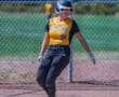 Softball player in yellow and black uniform stands on base, smiling, with helmet on, near chain-link fence.