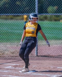 Softball player in yellow and black uniform stands on base, smiling, with helmet on, near chain-link fence.