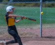 Softball player in action, hitting a pitch with a bat on a sunny day, wearing protective gear and team uniform.