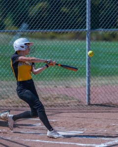 Softball player in action, hitting a pitch with a bat on a sunny day, wearing protective gear and team uniform.