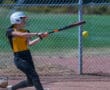 Softball player hitting a ball during a game, wearing yellow and black uniform, with a chain-link fence background.