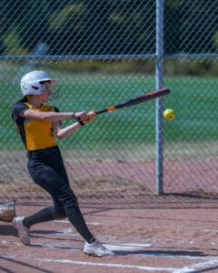 Softball player hitting a ball during a game, wearing yellow and black uniform, with a chain-link fence background.