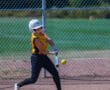 Softball player hitting a pitch during a game, wearing a yellow jersey and helmet, with a chain-link fence in the background.