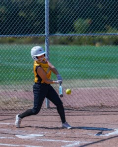 Softball player hitting a pitch during a game, wearing a yellow jersey and helmet, with a chain-link fence in the background.