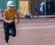 Softball player in action on the field, wearing yellow jersey and helmet, with dugout in background.