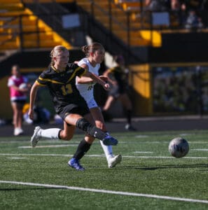 Soccer players in action on the field, one kicking the ball, wearing black and yellow jersey, daytime match.