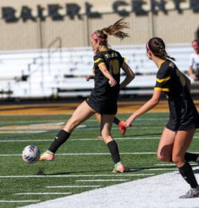 Soccer player kicking ball during game on field, wearing black and yellow jersey, with teammate nearby.