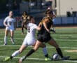 Two women competing fiercely in a soccer match, battling for control of the ball on a sunny field.