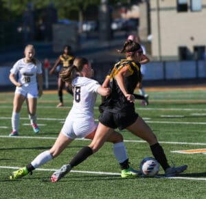 Two women competing fiercely in a soccer match, battling for control of the ball on a sunny field.