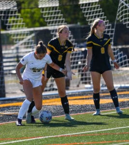 Girls playing competitive soccer on field, one in white and two in black, focusing on the ball.