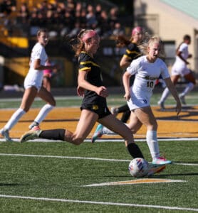 Female soccer player in black jersey dribbles ball past opponent on a sunny day.