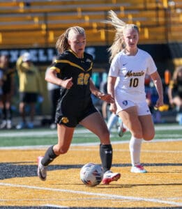 Two female soccer players in action, one in black and yellow, other in white, competing for the ball on the field.