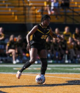 Soccer player dribbles ball on field during match, wearing black and yellow team uniform, focused on gameplay.
