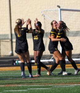 Soccer players celebrating a goal on the field, wearing black and yellow uniforms, under sunny weather.