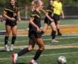 Female soccer player dribbling ball during match, teammates and referee in background.