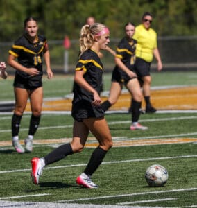 Female soccer player dribbling ball during match, teammates and referee in background.