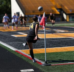 Soccer player in black jersey takes a corner kick during a match, with the ball in mid-air near the corner flag.