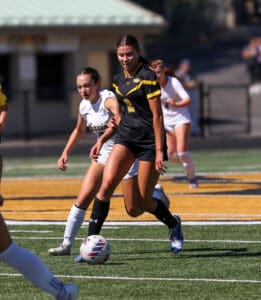 Girls' soccer match, player in black dribbles the ball past opponent on sunny field.
