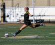 Soccer player kicks ball mid-air on field during sunny day, showcasing dynamic action and athleticism.