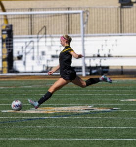 Soccer player kicks ball mid-air on field during sunny day, showcasing dynamic action and athleticism.