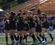 Women's soccer team celebrates on the field after a successful play, wearing black and yellow uniforms.