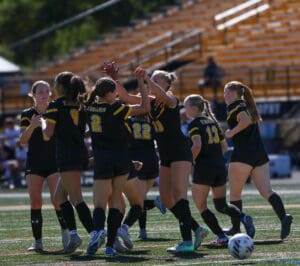 Women's soccer team celebrates on the field after a successful play, wearing black and yellow uniforms.