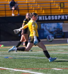 Soccer goalie kicks ball during match, wearing yellow jersey and gloves on grass field.