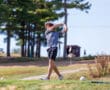Golfer swinging on a sunny day at a lush golf course with trees and a cart in the background.