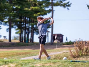 Golfer swinging on a sunny day at a lush golf course with trees and a cart in the background.