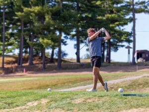 Golfer swinging club on green course, surrounded by tall trees on a sunny day.