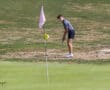 Golfer preparing to putt on a dry fairway near the green on a sunny day, with a pink flag in the foreground.