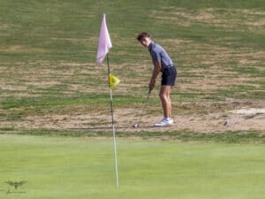 Golfer preparing to putt on a dry fairway near the green on a sunny day, with a pink flag in the foreground.