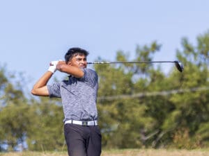 Golfer swings club on a sunny day, wearing a gray shirt and white gloves, with trees in the background.